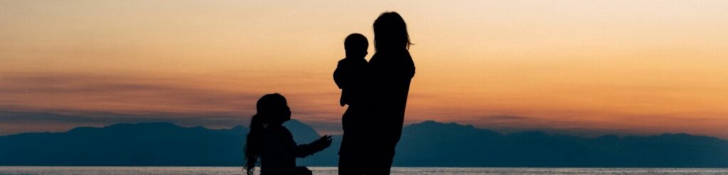 mulher segurando um bebê e uma menina pequena em frente ao pôr do sol com montanhas ao fundo