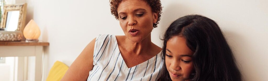 uma menina sentada na cama com sua mãe, e ambas lendo um livro juntas