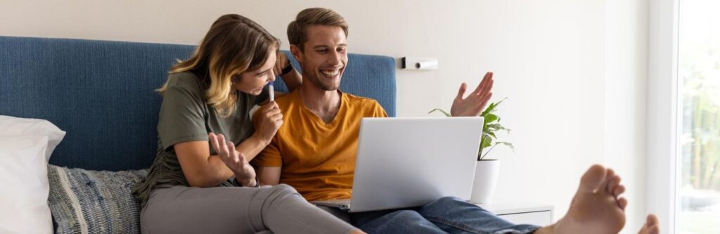 Casal sentado na cama rindo enquanto olha para um laptop.