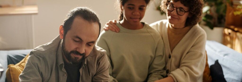 Um pai assinando um documento com sua filha e esposa ao fundo.